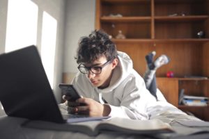 teenage boy staring at phone while a laptop is open in front of him and demonstrating how social media affects teens mental health