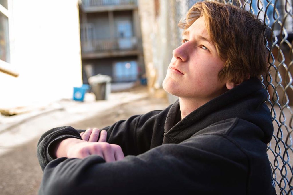 teenage boy sitting against chain link fence outdoors in an alley pondering the 7 tips to build confidence in teens