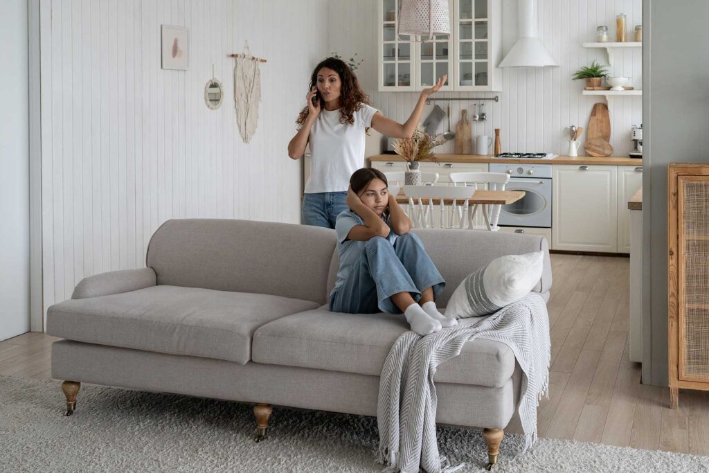 female teenager sitting on couch with back towards her mother who is on the phone talking to a therapist about how parents affect teenagers' mental health