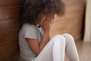 teenage girl sitting alone on the floor of her room with her back to the wall and her head buried in her hands experiencing the dangerous impact of childhood trauma and brain development