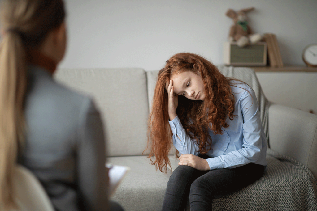 teenage girl seated on couch in therapist's office looking distraught and discussing how does teenage trauma affect adulthood