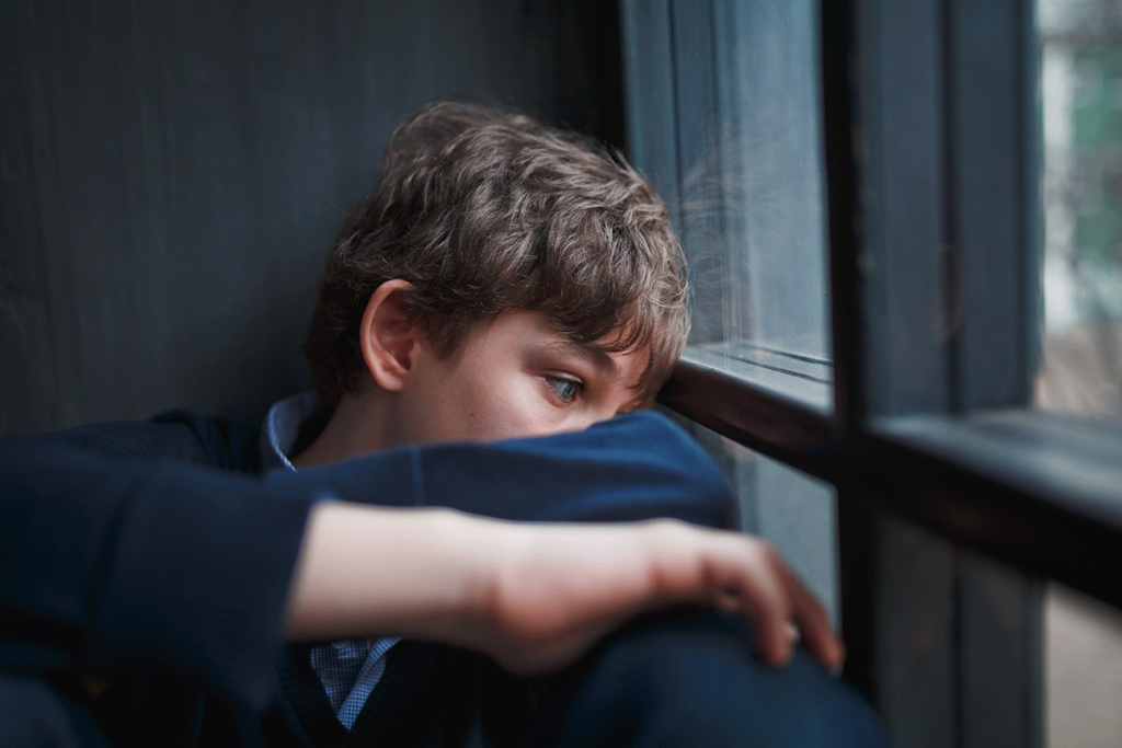 sad looking boy leaning dejectedly against window and wondering what are the effects of adolescent trauma