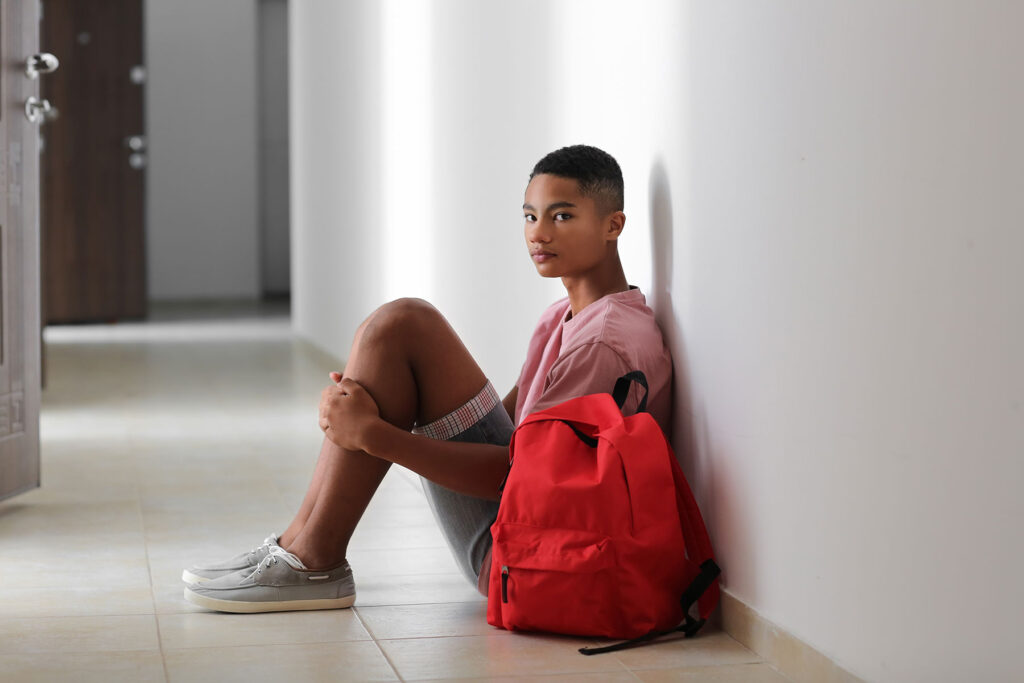 a teen sits on a floor with their legs to their chest near a backpack demonstrating how mental health affects high school students