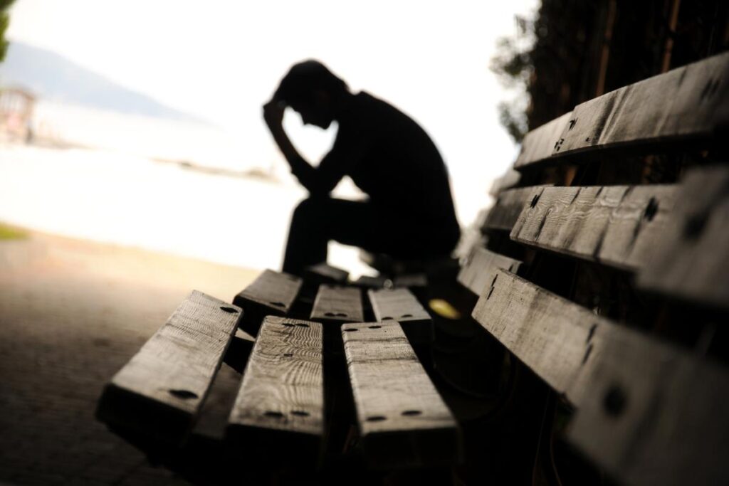 teenage boy seated alone on bench in remote area at sunset with his head bowed exhibiting one of 6 signs of co-occurring disorders in teens